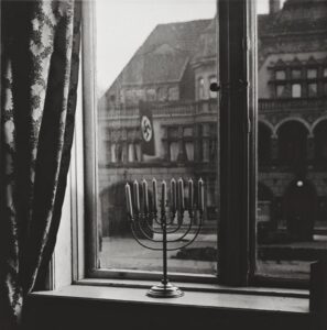 Photograph of a memorah in a window. A Nazi flag flies outside, 1931.
