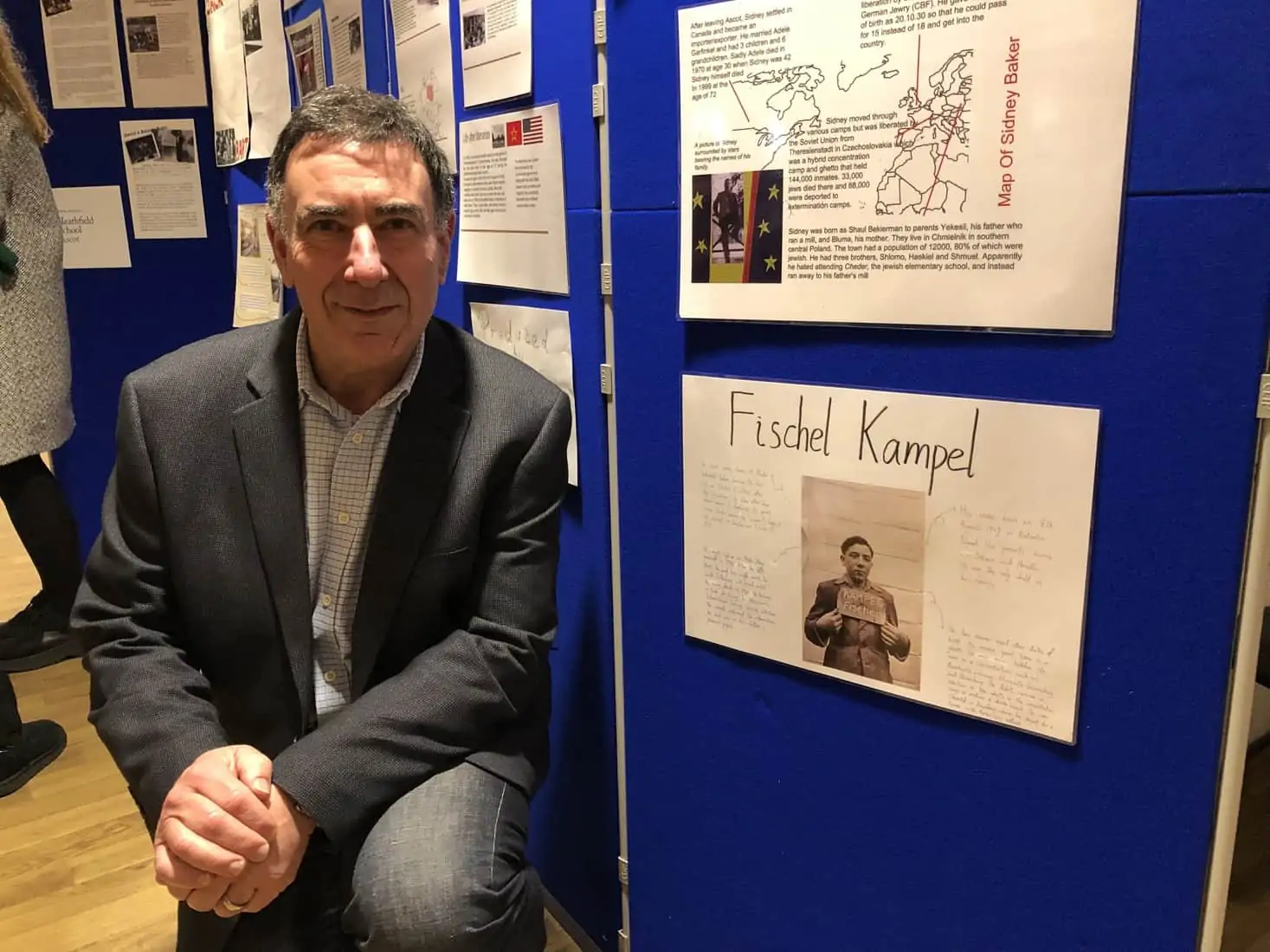 Photograph of Lawrence Kampel at the Ascot commemoration in January 2019. He poses next to a poster made by local school children about his father Fischel Kampel.